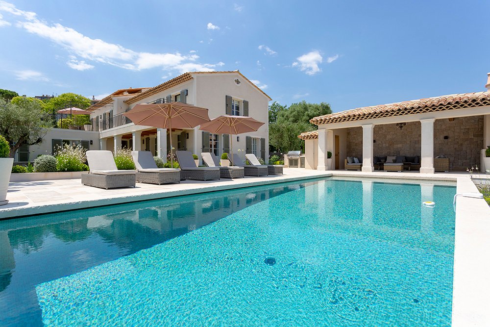 Luxury Mediterranean villa with terracotta roof tiles and light-colored stucco walls, surrounded by lush landscaping. A sparkling turquoise swimming pool is in the foreground, bordered by cushioned lounge chairs and large beige umbrellas. An outdoor covered seating area with stone walls completes the serene, sun-soaked setting.